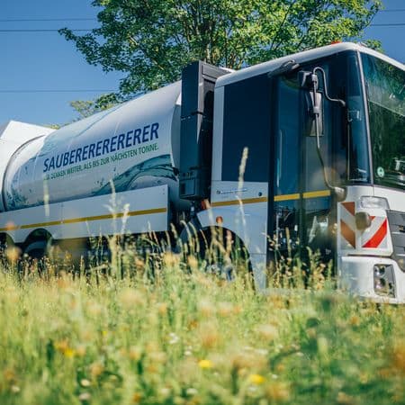 Ein Wasserstoff-Müllfahrzeug wird gezeigt, im Vordergrund Wiese, im Hintergrund blauer Himmel und ein Baum mit grünem Laub.