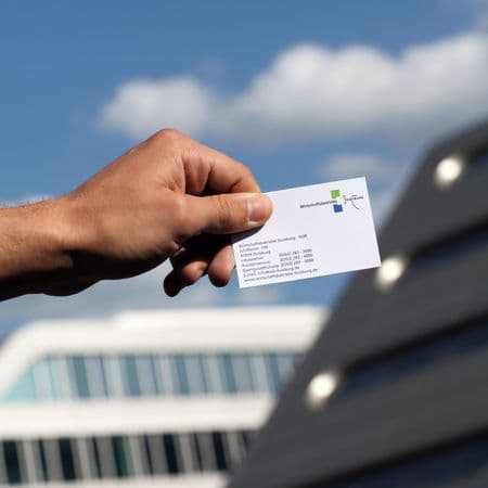 Gründung WBD-AöR in 2007: Hand hält eine Visitenkarte mit Logo WBD und Kontaktdaten. Im Hintergrund sieht man verschwommen das H2-Office im Duisburger Innenhafen.