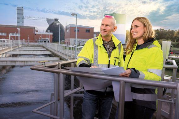 Eine Mitarbeiterin und ein Mitarbeiter in gelber Arbeitsjacke schauen über eine Kläranlage und halten einen Plan in den Händen