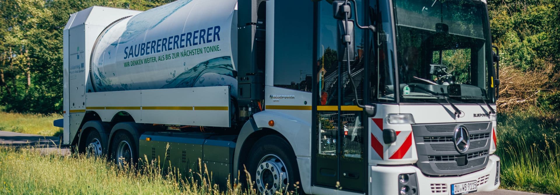 Müllfahrzeug Weißes Müllfahrzeug auf Straße, vorne auf dem Bild Wiese, hinten Bäume und blauer Himmel
