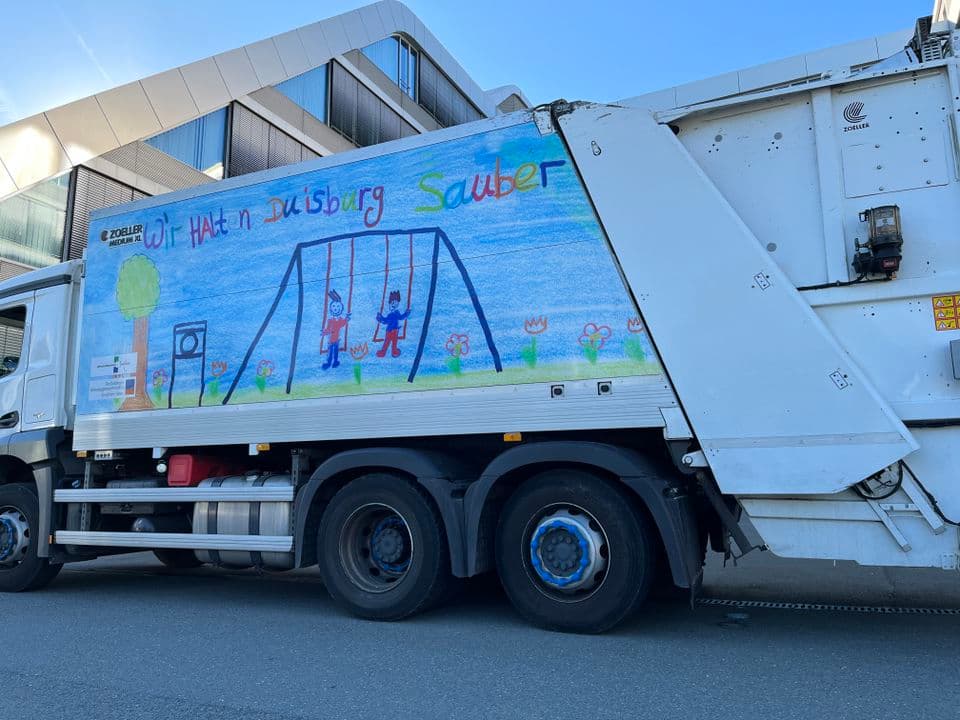 WeißesMüllfahrzeug mit Bemalung von Kindern. Man sieht einen gemalten Spielplatz.