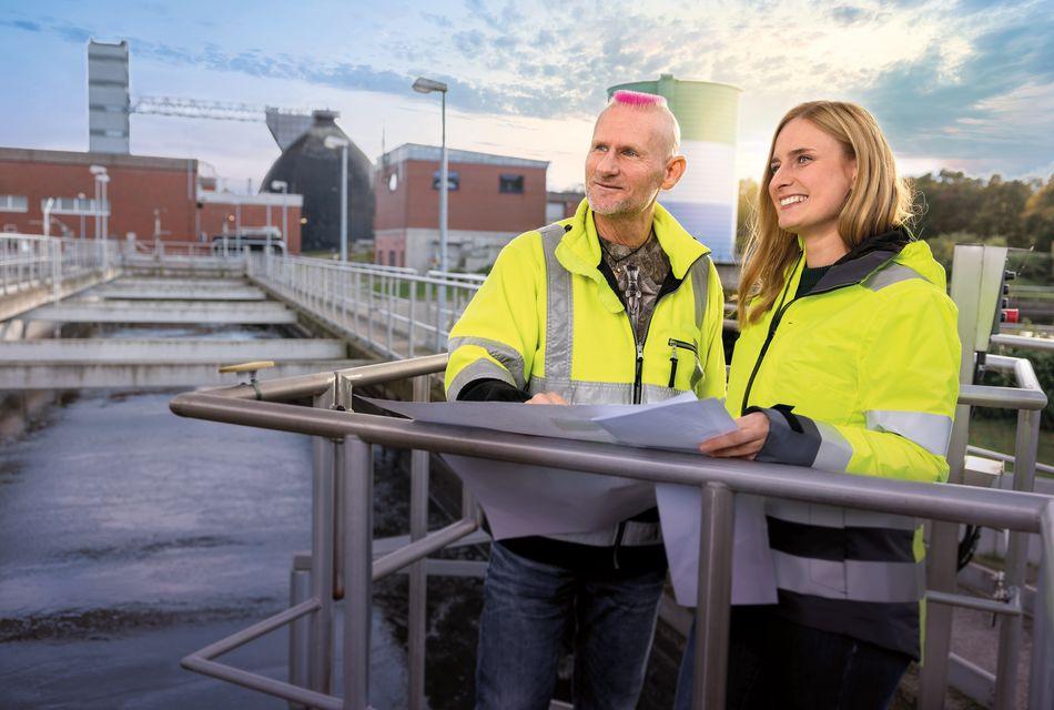 Motiv Kläranlage Eine Mitarbeiterin und ein Mitarbeiter in gelber Arbeitsjacke schauen über eine Kläranlage und halten einen Plan in den Händen